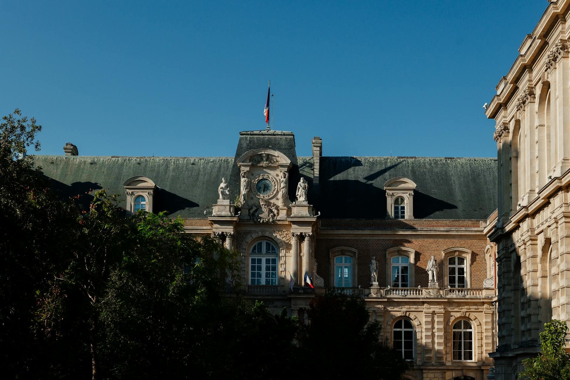 Elegant facade of a historic French building with statues and French flag on a sunny day.